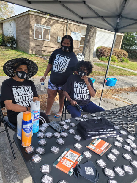 Three women are gathered together under a canopy, all of them are wearing matching shirts that say Black Voters Matter. There are buttons, shirts, face masks, and wrist bracelets spread on the table in front of them.