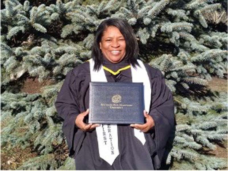 Debra Jackson smiles while wearing her graduation cap and gown and holding her diploma in front of her. 
