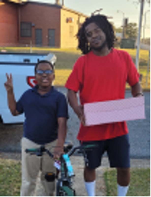 A young man holding a box and a little boy on a bike stand together. 