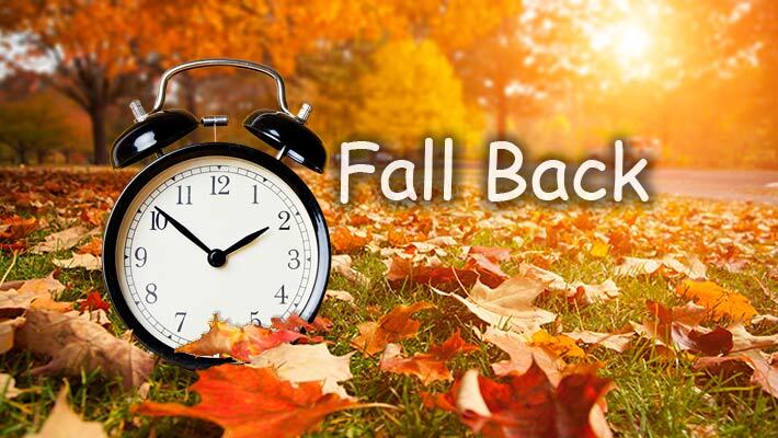 Alarm clock laying on autumn leaves with text that reads: Fall Back.
