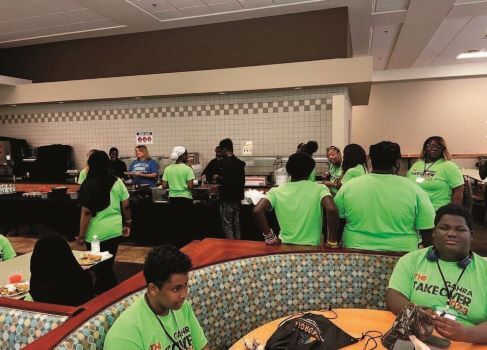 A cafeteria with students in line, at tables and booths. 