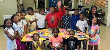 Group of 15 kids and a teacher stand around a table and smile for a picture.