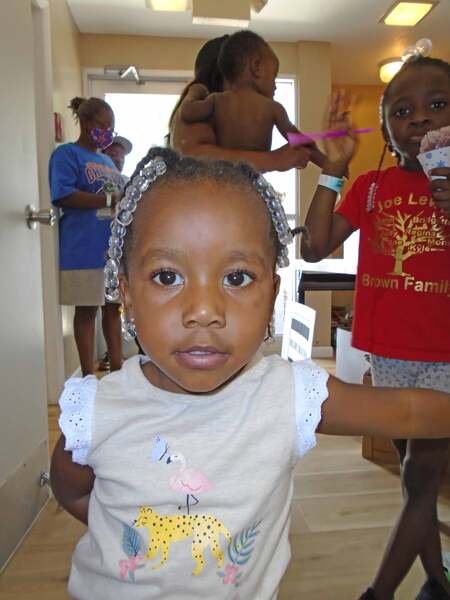 A little girl is staring into the camera, caught off guard by the flash; the girl behind looks on and waves as she eats a snowcone. 