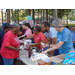 A volunteer behind the ice-cream table extends a chocolate chip cookie out to a resident who is holding a cup of ice-cream in her hand. 