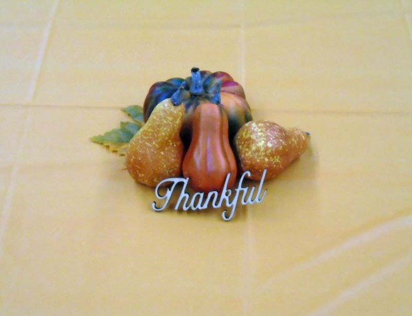 A pumpkin and some gourds with the word Thankful in front sit in the middle of a table to create a center piece