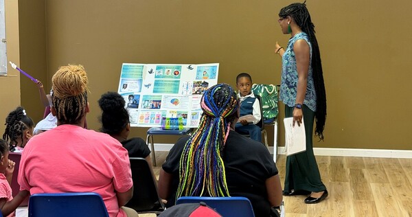A woman describing a informational poster board to a group of people.